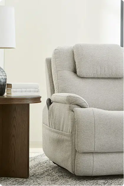A close-up of a beige power lift recliner chair with plush cushioning and a remote tucked into the side pocket, positioned next to a dark wood side table with a lamp and stacked books.