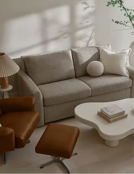A compact beige sofa bed is styled in a warm, sunlit room with modern brown accent chairs, a sculptural white coffee table, and minimalist decor. Neutral throw pillows and soft shadows add to the cozy, contemporary vibe.