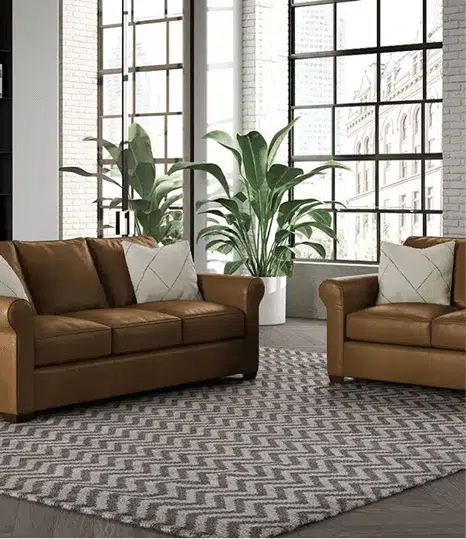 A matching set of caramel leather sofas with rolled arms is arranged in front of large industrial-style windows. The living room features geometric throw pillows, tall green plants, and a bold zigzag area rug on dark hardwood floors.