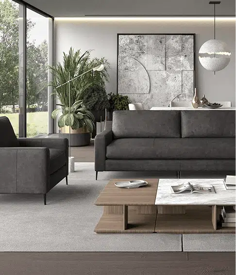 A sleek charcoal gray sectional and matching armchair sit in a contemporary living room with floor-to-ceiling windows and minimalist decor. A low wood and marble coffee table anchors the space, complemented by large plants, modern art, and soft ambient lighting.