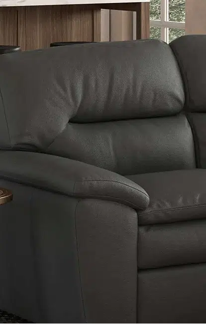 A close-up of a dark charcoal leather sofa shows its plush armrest, supportive back cushion, and smooth texture. The edge of a round wood side table is visible beside it, adding a touch of warmth to the modern design.