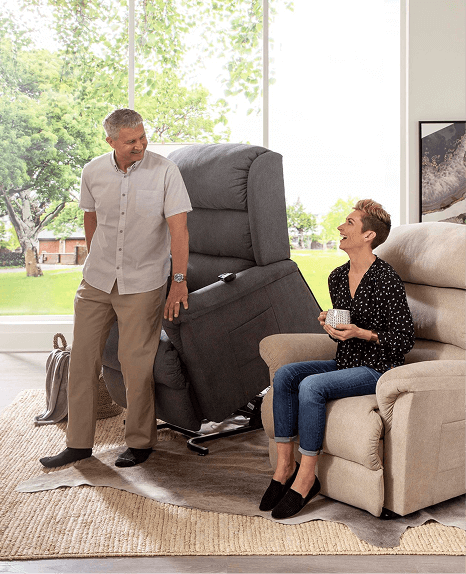 A smiling man stands next to a dark gray power lift recliner in the raised position, while a woman holding a teacup sits on a beige recliner beside him. They appear to be chatting in a bright living room with large windows overlooking a green yard.