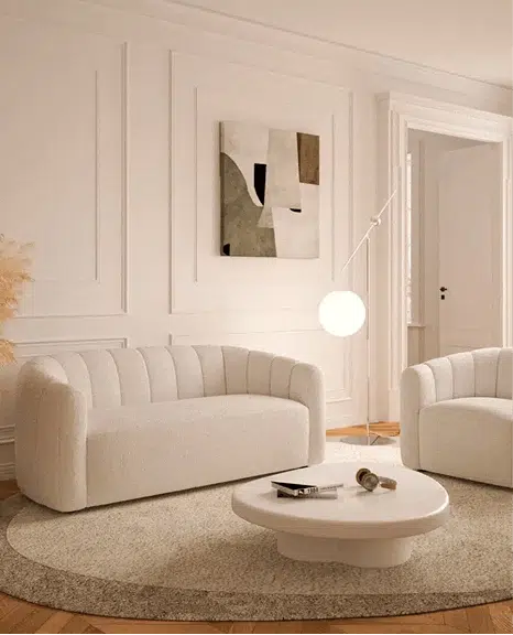 A cream-colored curved sofa with vertical tufting is paired with a matching chair in an elegant room with paneled walls. A round white coffee table, sculptural floor lamp, and abstract wall art complete the soft, minimalist aesthetic.