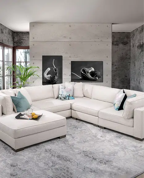 A large cream-colored sectional with a chaise lounge is styled in a modern living room with a textured gray accent wall and black abstract art. Colorful throw pillows and a tray with drinks add comfort and contrast to the light, elegant space.