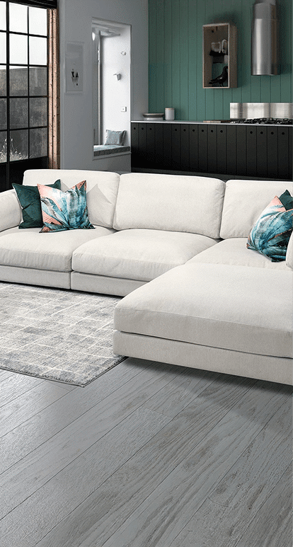 A modern cream-colored sectional with a chaise lounge is set against dark cabinetry and a green accent wall in an open-concept living space. Colorful tropical throw pillows add contrast, and the light gray wood flooring complements the neutral tones.