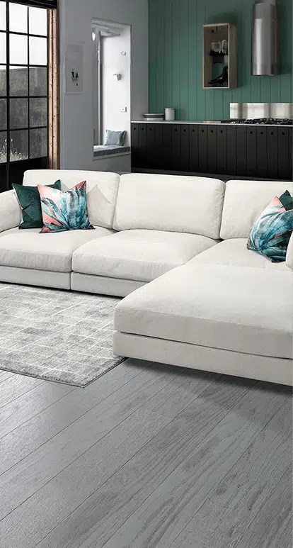 A modern cream-colored sectional with a chaise lounge is set against dark cabinetry and a green accent wall in an open-concept living space. Colorful tropical throw pillows add contrast, and the light gray wood flooring complements the neutral tones.