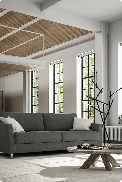 A dark gray modern sofa bed sits in a bright, loft-style room with tall industrial windows and minimalist decor. A low wood coffee table holds a book and eyeglasses, while angled ceiling beams and natural light create an airy, open feel.