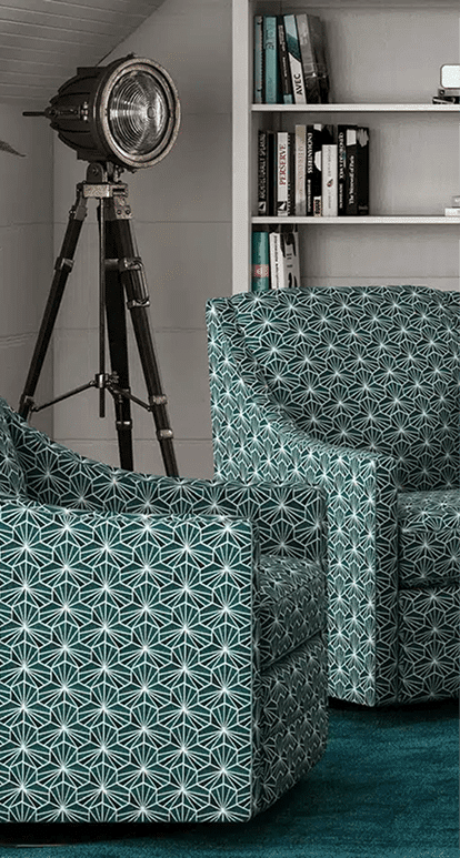 Two patterned swivel chairs with a bold geometric teal and white design are positioned in a cozy reading nook with a deep teal carpet. A vintage-style spotlight floor lamp and white built-in bookshelf filled with books complete the eclectic space.