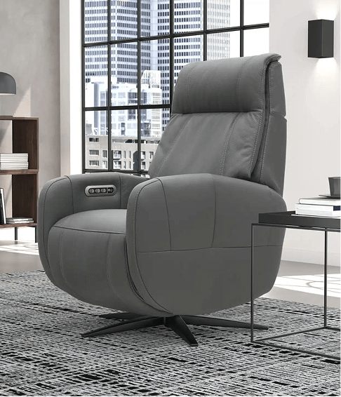 A modern gray leather swivel recliner with built-in power controls sits in a stylish high-rise apartment. It's placed on a patterned rug next to a black metal side table, with large windows showcasing a city skyline.