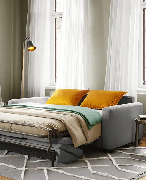 A gray sofa bed is fully extended with a neatly made mattress, tan bedding, and vibrant yellow pillows in a softly lit room with sheer white curtains. A black and gold floor lamp and geometric rug complete the cozy, functional setup.