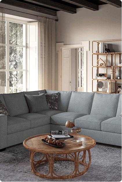 A spacious light gray sectional sofa is arranged in an L-shape in a cozy living room with tall windows, exposed wood beams, and a rattan coffee table topped with books and snacks. A bamboo-style bookshelf adds a warm, natural touch to the space.