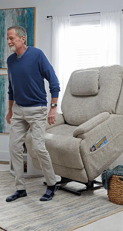 An older man in a blue sweater and light pants uses a beige power lift recliner to assist in standing up. The chair is angled forward in lift mode and has a promotional flyer in its side pocket. Sunlight filters through white curtains in the background.