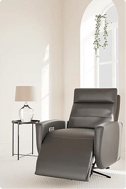 A sleek gray leather recliner in a partially reclined position sits in a minimalist room with soft natural light. Beside it is a small glass lamp on a modern black metal side table, with an arched window and hanging greenery in the background.