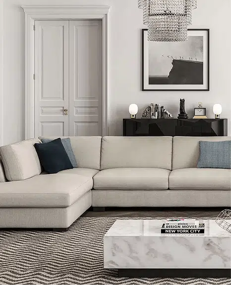 A neutral-toned sectional with a chaise sits in a sophisticated living room featuring a marble coffee table, zigzag-patterned rug, and black console table topped with modern decor. A framed monochrome print and crystal chandelier add upscale elegance.