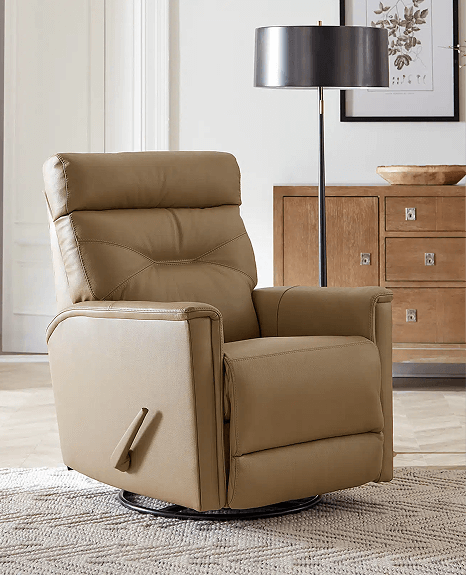 A tan leather manual recliner with a side lever is featured in a bright, tastefully decorated room with a floor lamp, wood dresser, and framed artwork. The chair sits on a neutral woven rug, offering a cozy, classic feel.