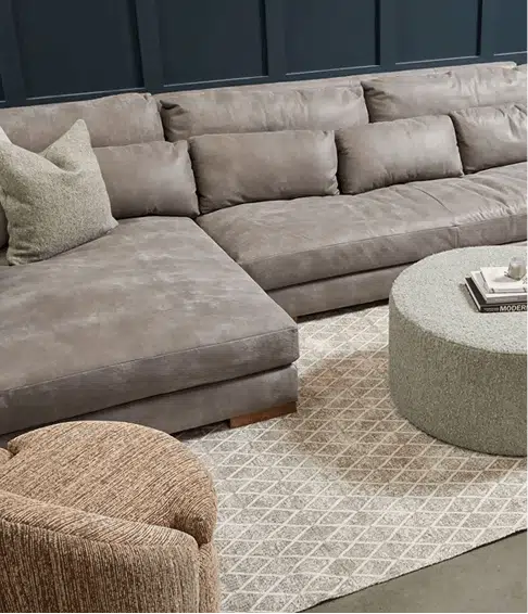 A large taupe suede sectional with deep seats and a chaise is styled in a cozy living room with a dark blue accent wall. A round textured coffee table holds books, and a patterned rug and woven chair add warmth and texture to the space.