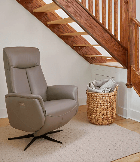A sleek taupe leather swivel recliner with a slim profile is positioned under a wooden staircase on a light rug. A woven basket filled with folded blankets sits nearby, adding a cozy touch to the minimalist corner.