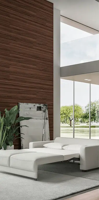 A white leather sofa bed is extended in a bright, modern living room with floor-to-ceiling windows and a warm wood accent wall. A large green plant and a leaning abstract canvas add texture to the sleek, minimalist space.